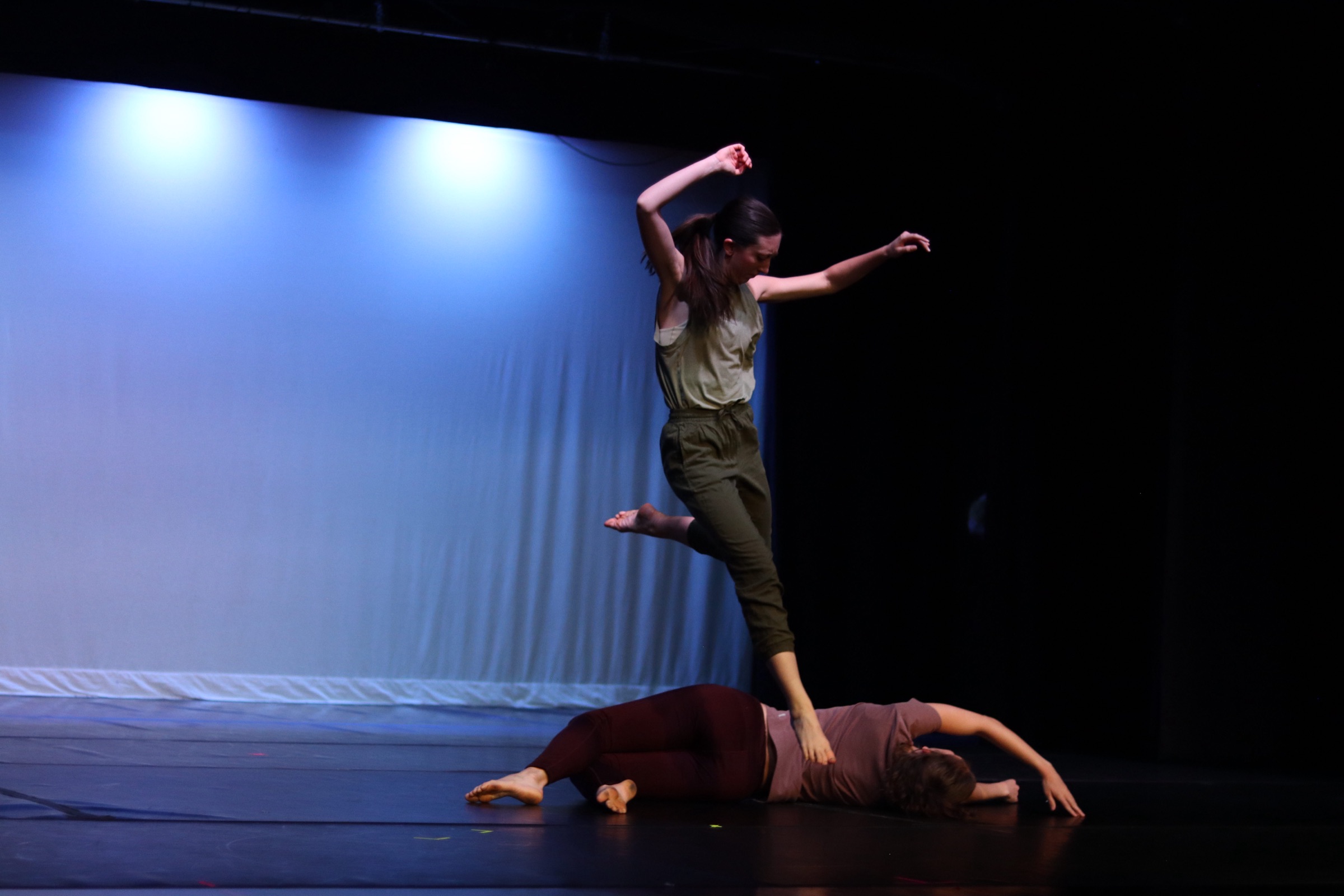 A dancer leaps mid-air above a partner lying on a darkened stage