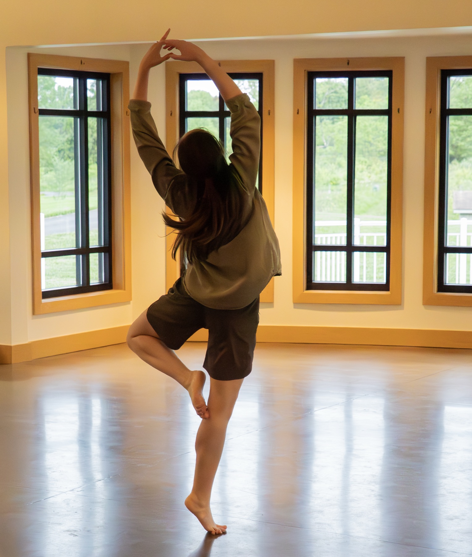 A dancer in a sunlit studio with arms overhead and one leg raised